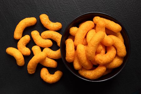 Extruded cheese puffs in a black ceramic bowl next to spilled cheese puffs isolated on black. Top view.の写真素材