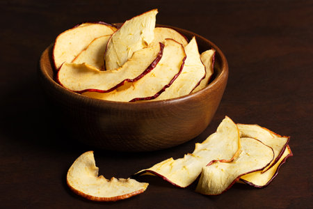 Dried apple slices in a brown wood bowl next to spilled dried apple slices on dark wood background.