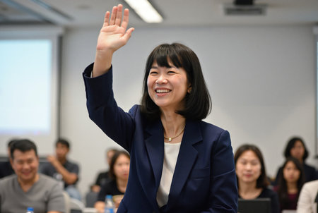 Asian woman with a bob haircut, smiling warmly while raising her right hand in a classroom setting. She wears a grey blazer over a white blouse. In the background, a projector screen displays a presentation, and other students are seated, slightly blurred.の素材