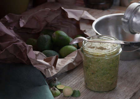 Fruits of feijoa in rough paper and a jar of feijoa jam with green cloth, grinder and metal bowl on wooden table in vintage kitchenの写真素材