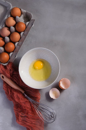 A box with brown eggs, a bowl with yolk and a whisk on gray kitchen table.の写真素材