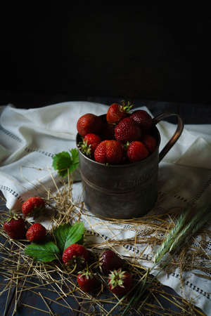 Ripe freshly picked up strawberry in metal rustic cup on white towel with straw and berries, low key, selective focusの写真素材