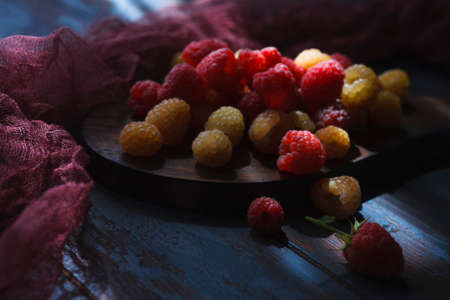 Red and yellow raspberry on wooden board with dark red cloth, low key, close up