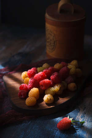 Red and yellow raspberry on wooden board with wooden pot in the background, low keyの写真素材