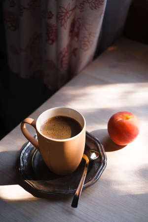 A cup of coffee on vintage matal plate on white wooden table with a peach at morning light in the interior, summer morning concept.の写真素材