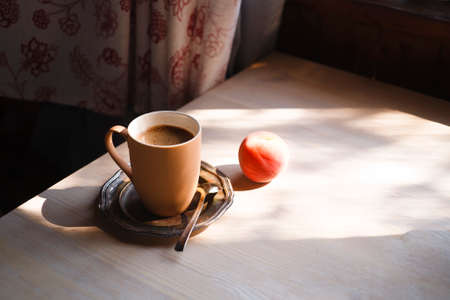 A cup of coffee on vintage matal plate on white wooden table with a peach at morning light in the rustic kitchen, summer morning concept.の写真素材