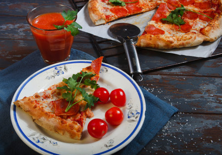 Lunch break with traditional pepperoni pizza on blue wooden table with a slice of pizza on white plate with cherry tomatoes and a glass of tomato juice.の写真素材