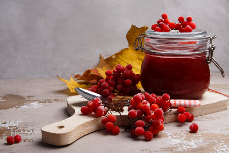 A glass jar with viburnum fruit jam and ripe red viburnum berries on gray background, autumn...