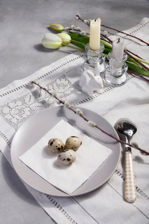 Table Easter decor set with quail eggs on plate, willow branches, white tulip and candles on white embroidered table cloth, selective focus.の写真素材