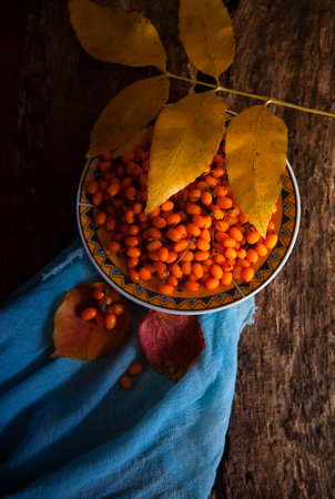 Sea-buckthorn berries on plate on a wooden background with blue cloth and yellow autumn leaf, flat layの写真素材