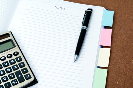 Blank notebook page with black ballpen, calculator and multi colored post it on wooden backgroundの写真素材