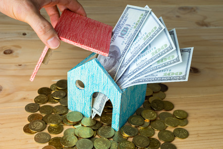 Conceptual image of small house with gold coins and hundred dollar bills on wooden surface. Selective focus.の写真素材
