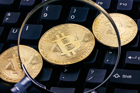 Close-up shot of golden bitcoin cryptocurrency through a magnifying glass on top of computer keyboardの写真素材