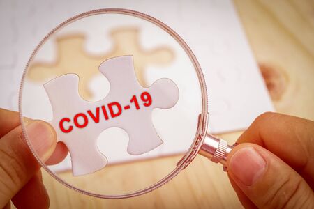 Hand with magnifying glass searching for missing piece of jigsaw puzzle with Covid-19 word.の写真素材