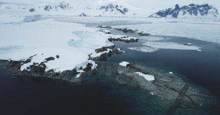 Snow covered rocky coastline in Antarctica. Aerial view frozen polar ocean coast bay with rocks underwater in cold water, mountains in background. Antarctic travel and exploration. Aerial drone shotの写真素材