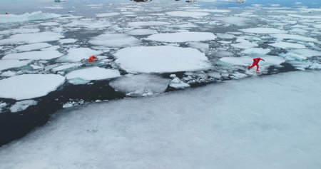 People in Antarctica ocean frozen ice floes. Two man in orange immersion suits run melting ice,...