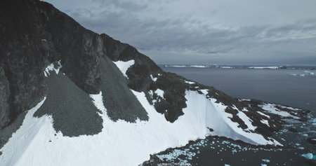 Snow covered dark mountain slope on Antarctica ocean coast. Dramatic landscape in moody weather day....