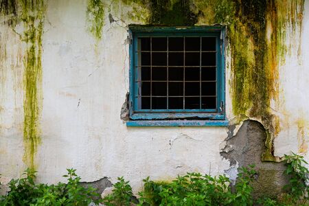 Broken blue window with metal frame on a damaged white wall of an abandoned building.