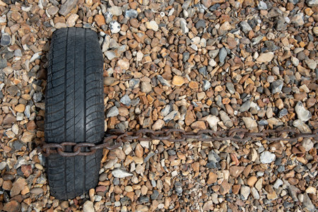 Rusty chain fastened on an old black vehicle wheel. Beach gravel background. Boat moored in the shoreの写真素材