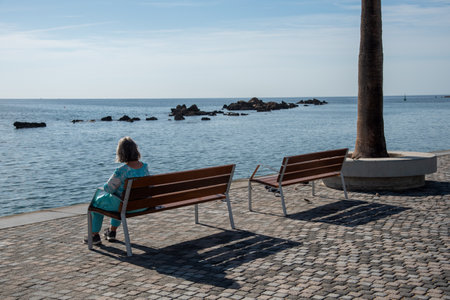 Elder woman siting alone on a wooden bench overlooking and enjoying seaの写真素材