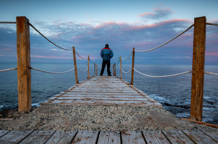 Person standing at the edge of a wooden pier in the sea at sunrise . People active outdoors enjoy oceanの写真素材