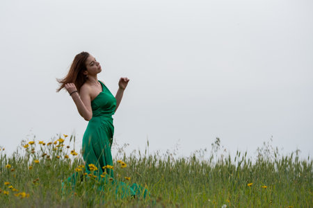 Young woman holding hair wearing a green long dress walking in blooming rural countryside field outdoorsの写真素材