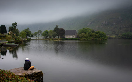 Tourist relaxing by gougane barra lake with saint finbarrs oratory in background on a foggy day