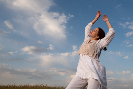 Young woman wearing white dress dancing outdoors against blue cloudy sky.の写真素材