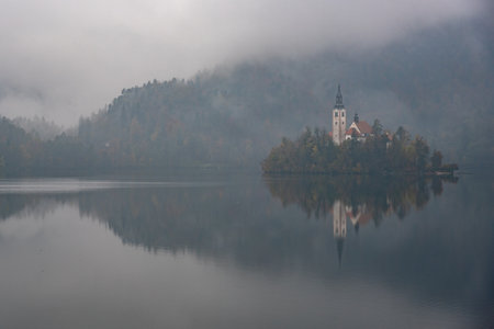 Assumption of mary pilgrimage church Lake bled island reflecting on calm water during foggy autumn dayの写真素材