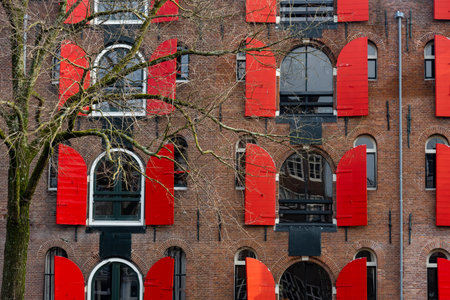 Partially opened red window shutters create a vibrant contrast against the brown brick facade of a typical amsterdam buildingの写真素材