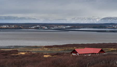 Red-roofed cabin sits among low vegetation by a calm lake, with snowy mountains visible in the...