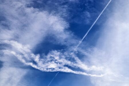 Aircraft condensation contrails in the blue sky inbetween some cloudsの写真素材