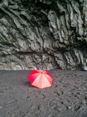 A lone red umbrella on the endless black beaches of Iceland in front of black basalt rock formationsの写真素材