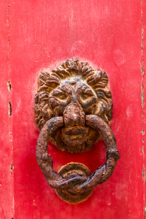 Close-up of an old rusty lion head door knocker on a vibrant red wooden door, adding a touch of...
