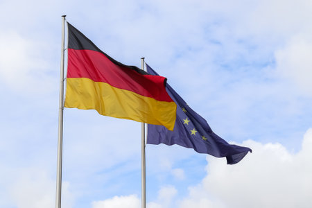 Flags of Germany and European Union waving on flagpoles against blue sky with some clouds