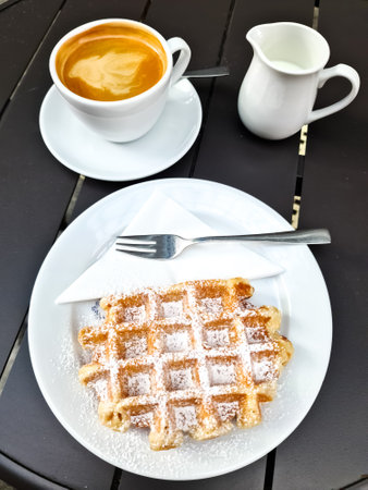 Tasty waffle with powdered sugar served on a white plate with coffee and milk jug in a cafeの写真素材