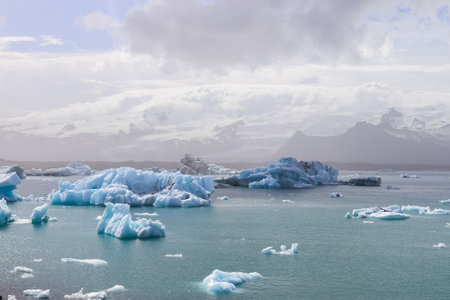 Icebergs are floating in Jokulsarlon glacier lagoon with a view of snow capped mountains and cloudy...