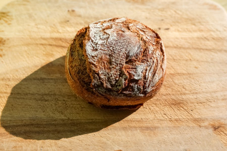 Freshly baked round sourdough bread loaf resting on a wooden cutting board, casting a strong shadow...