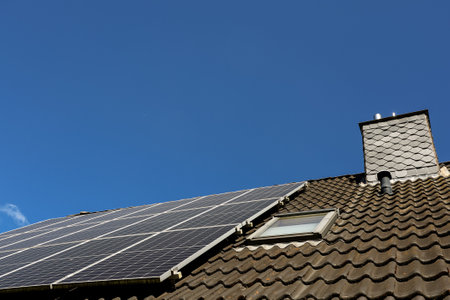 Photovoltaic panels are installed on a rooftop with a skylight and chimney against a clear blue sky,...