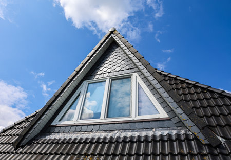 Modern dormer window integrated into a slate tiled roof, reflecting the blue sky and white clouds