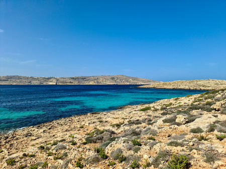 Turquoise water gently lapping against the rocky coast of Comino island, with the larger island of...
