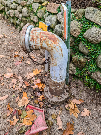 Rusty industrial pipe with a chain and bolts emerging from the ground, surrounded by colorful autumn oak leaves, contrasting with a weathered stone wall and green ivyの写真素材