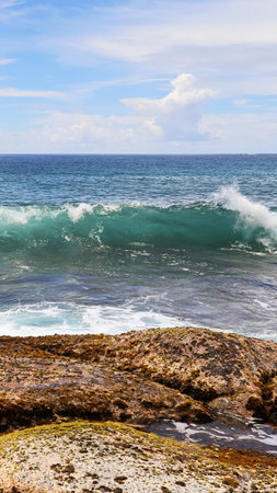Ocean wave breaking with white foam against the rocky coastline of a tropical island, featuring...