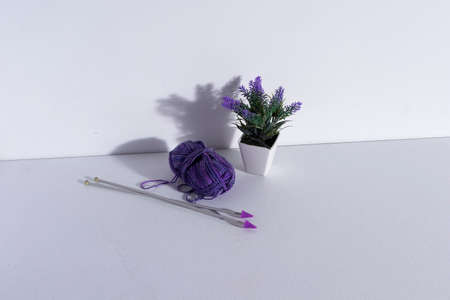 Artificial plant with purple flowers and white pot, next to a ball of purple wool with knitting...