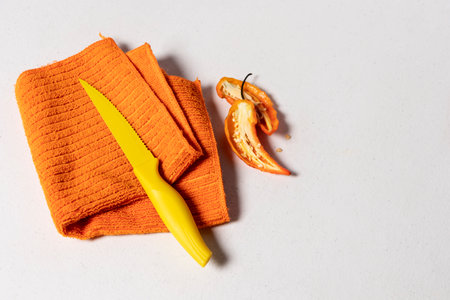 Orange dishcloth folded with a small yellow knife next to a pepper on top on a white backgroundの写真素材