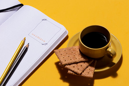 Yellow cup with coffee and cookies next to bullet journal notebook with tools to plan the goals of...