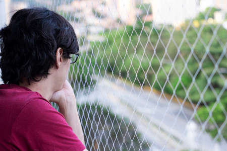 Black haired man and red t-shirt pensive on balcony of building with security mesh for pets