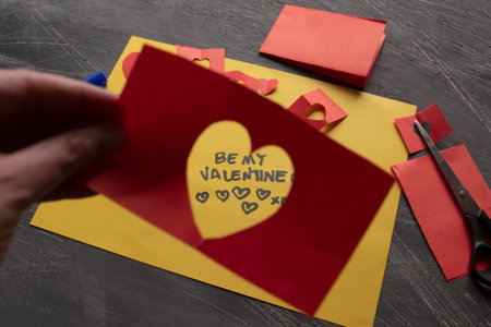Kid-Style Valentine Card Creation with Blue Ink and Red Scraps, A charming flat lay of a yellow paper note featuring the handwritten blue text Be My Valentine, surrounded by irregular red paper heart cutouts, perfect for DIY holiday concepts and cards.