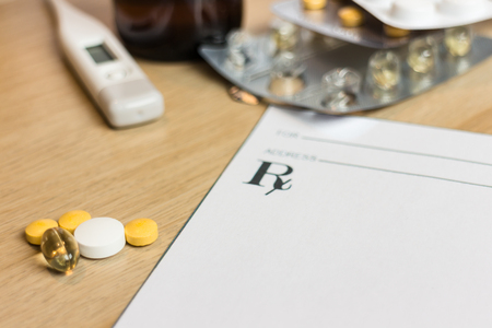 Varied pills next to blank prescription and thermometer and syrup in the backgroundの写真素材