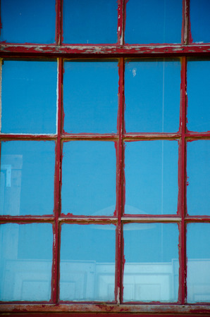 Window in old building  with faded and chipped red paint on pane that creates geometric pattern and reflects blue sky.の写真素材
